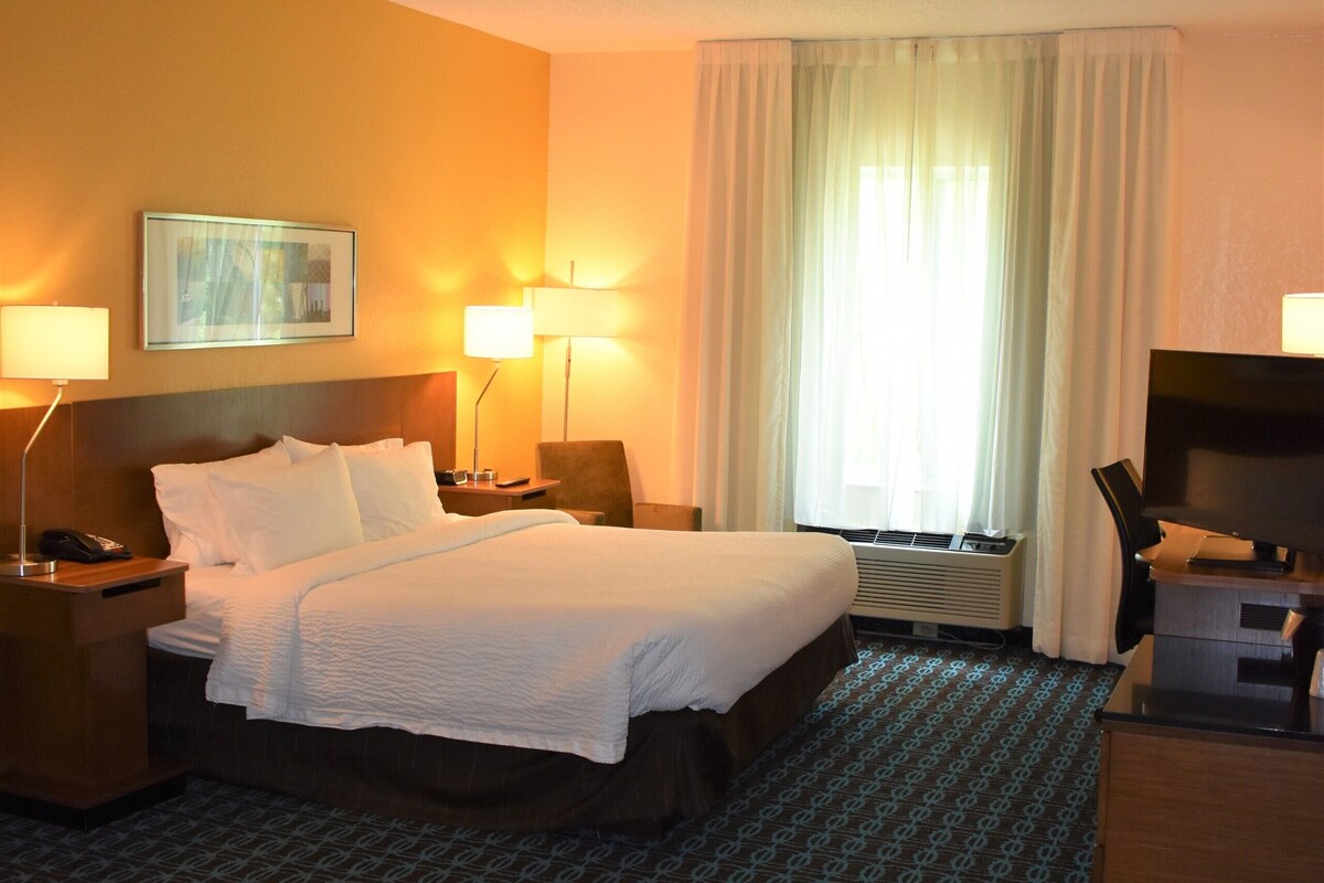 A hotel room with a bed, two bedside tables with lamps, a desk with a chair, a television, and a window with curtains.