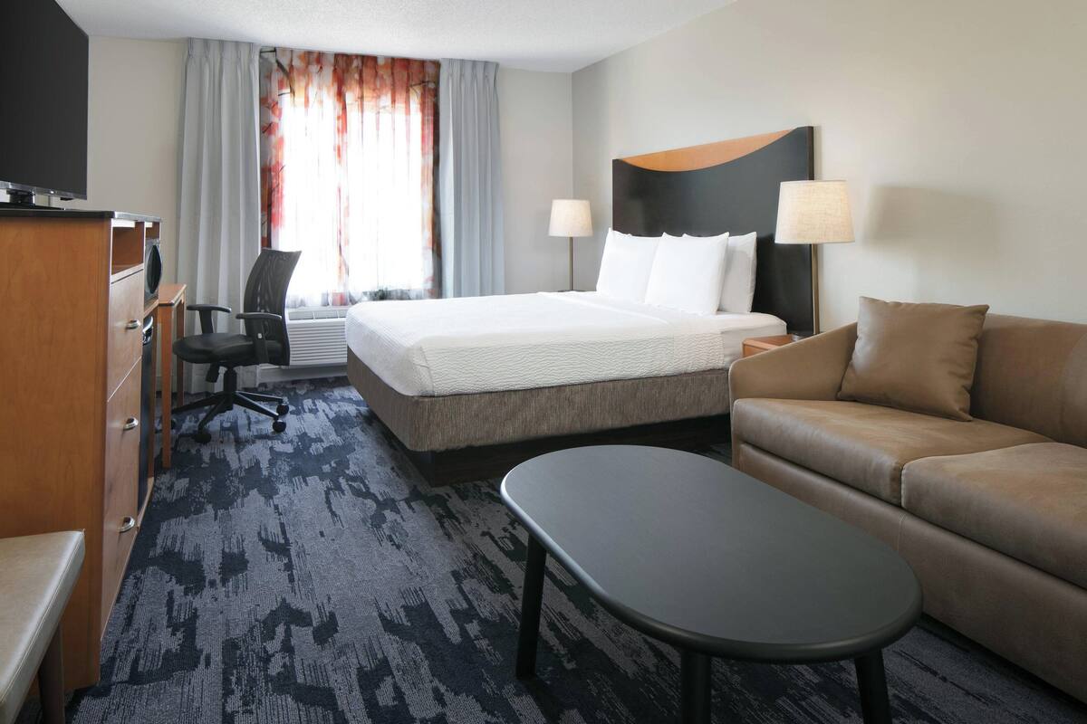 A hotel room with a bed, a sofa, a desk with a chair, a TV, and a window with curtains.