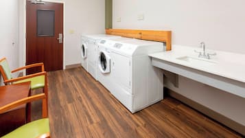 A laundry room with a wooden floor, a washing machine, a dryer, a sink, and a wooden chair.