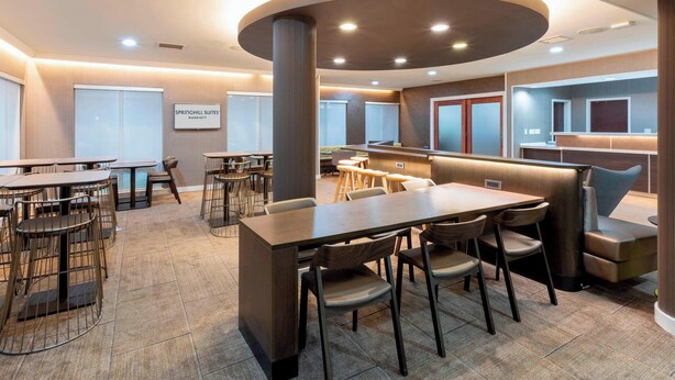 A modern hotel lobby with multiple seating areas, including a central dining table with chairs, a bar area, and a seating section with cushioned chairs.