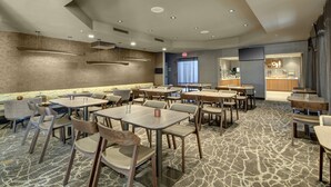 A modern hotel dining area with multiple tables and chairs, a patterned carpet, and a kitchen in the background. - SpringHill Suites Port St. Lucie (Port Saint Lucie)