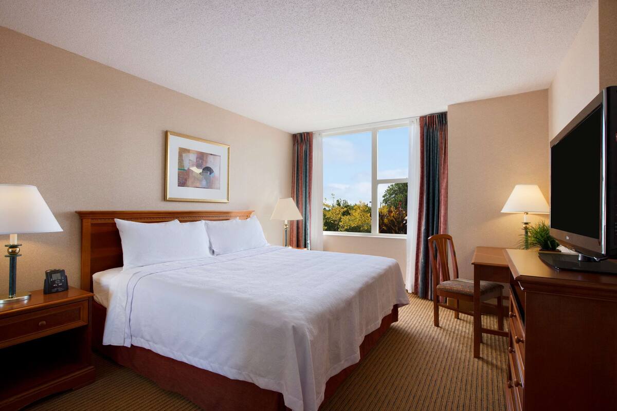 A hotel room with a bed, bedside tables, a desk, a television, and a window with curtains.