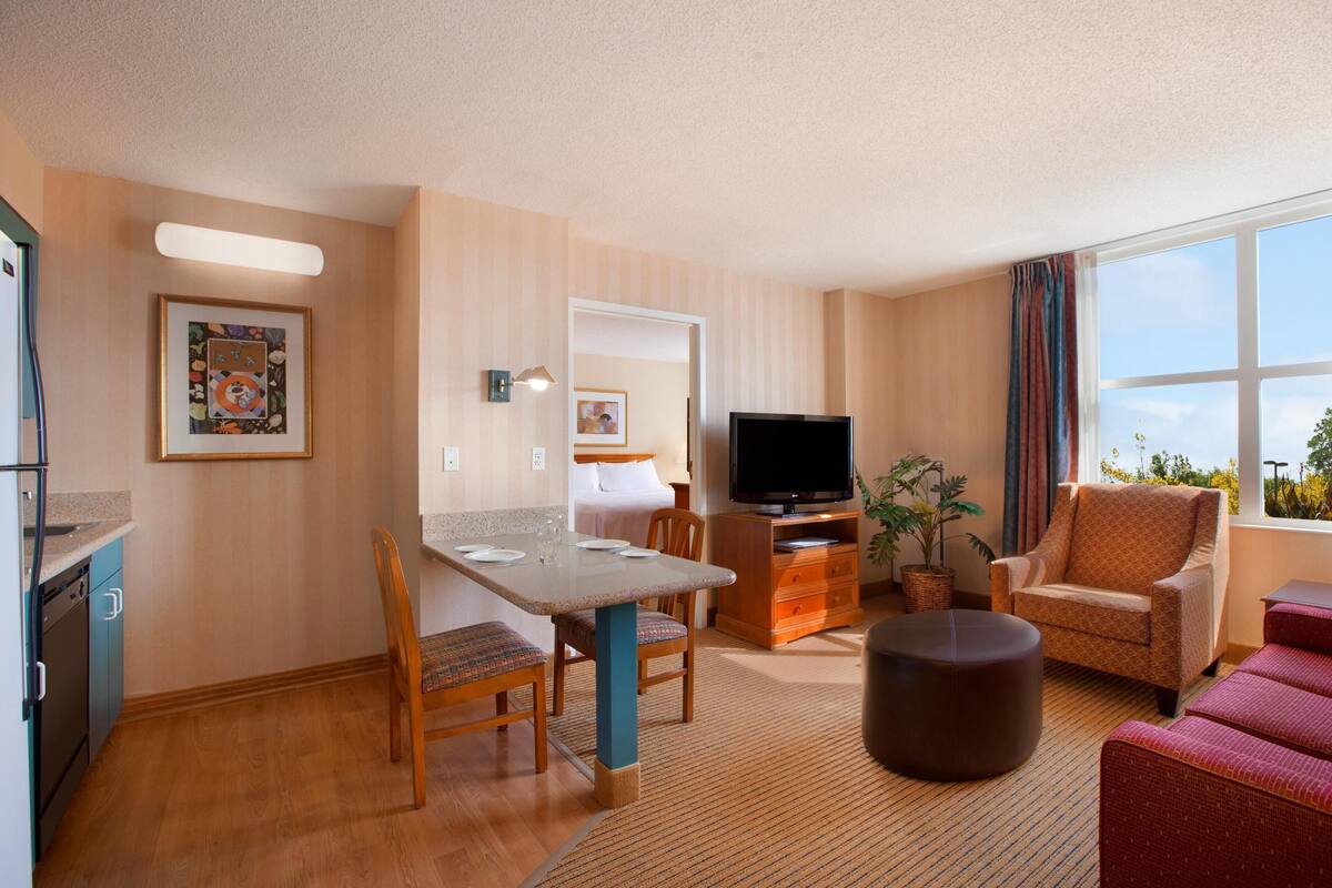 A hotel room with a dining area, a TV, a sofa, and a window with a view.