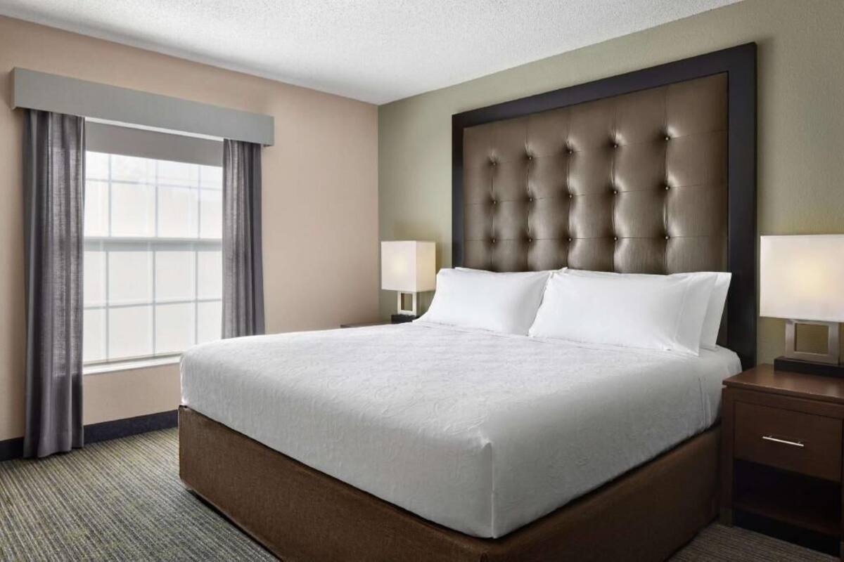 Une chambre d’hôtel avec un grand lit, deux tables de chevet avec des lampes, une fenêtre avec des rideaux et une tête de lit capitonnée.