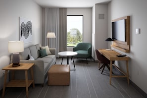 A modern hotel room with a sofa, a round table, a green armchair, a TV, and a window with curtains.