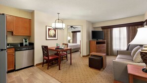 A hotel room with a kitchenette, a dining area with a table and chairs, a sofa, a TV, and a lamp. - Homewood Suites Dallas/Park Central (Dallas)