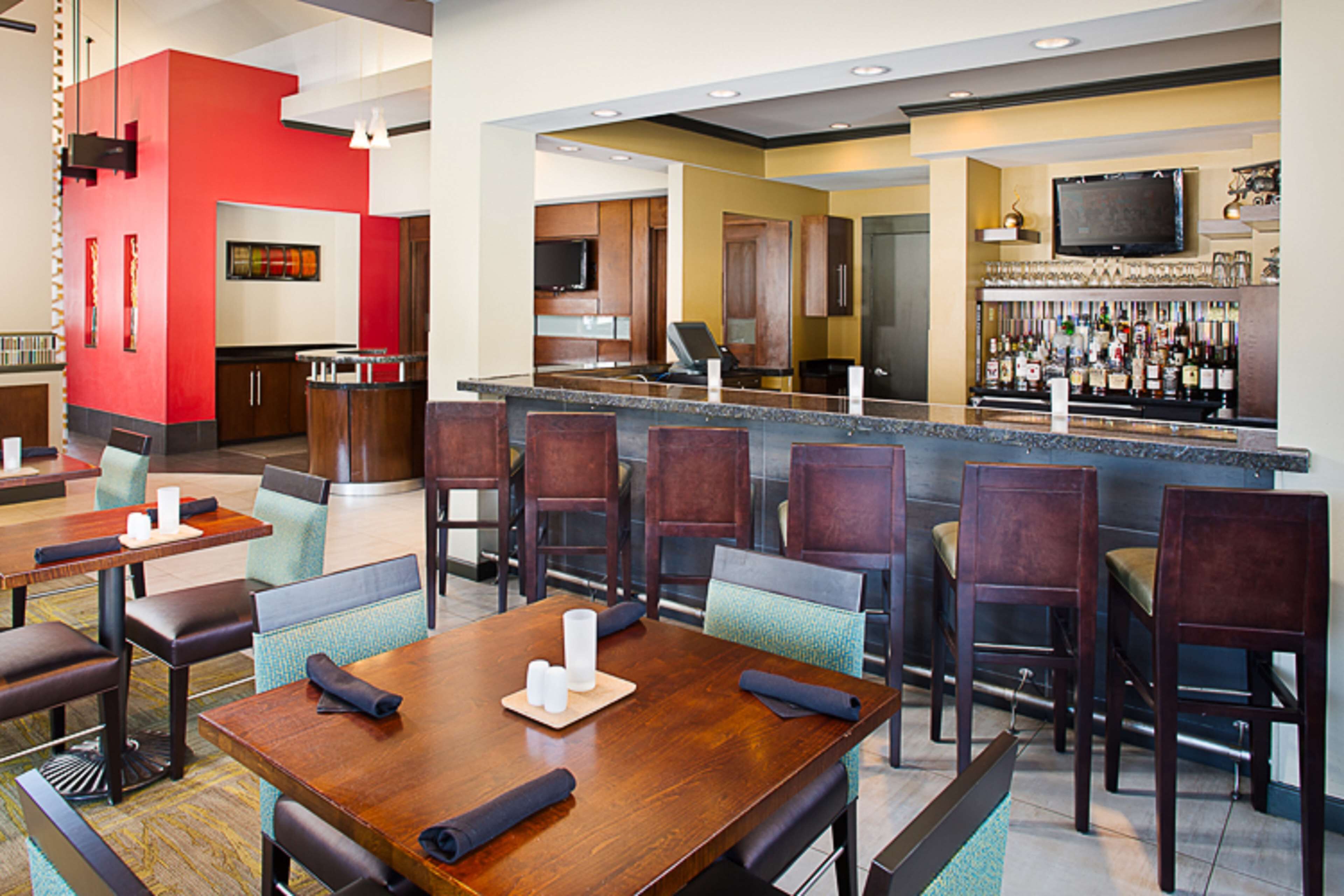 A hotel lobby with a bar area, dining tables, and a kitchen in the background.