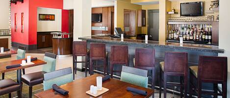 A hotel lobby with a bar area, dining tables, and a kitchen in the background.