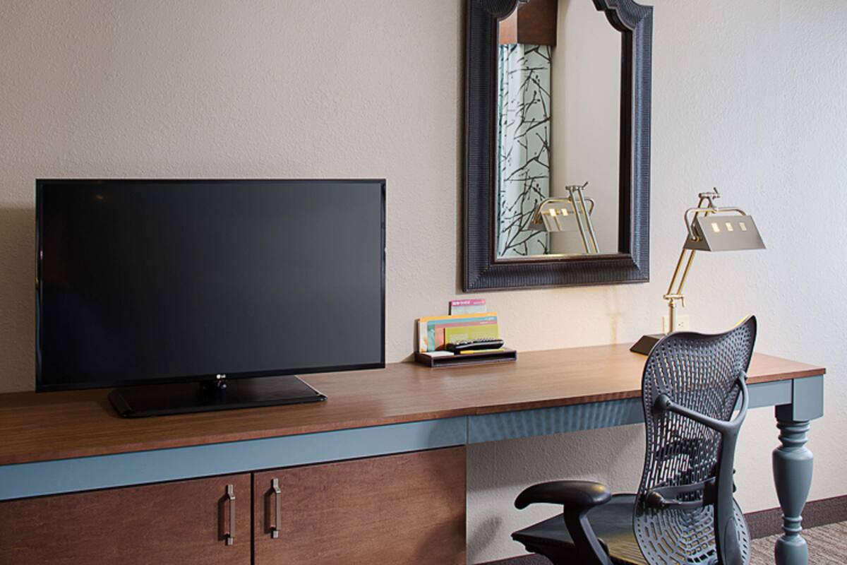 Habitación de hotel con un escritorio de madera, un televisor de pantalla plana, una silla, una lámpara y un espejo.