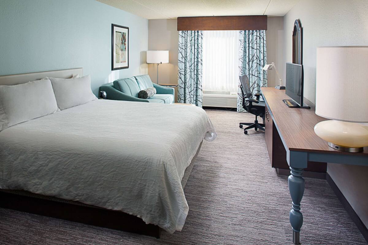 Habitación de hotel con una cama grande, un escritorio con computadora, una silla y un ventanal con cortinas.