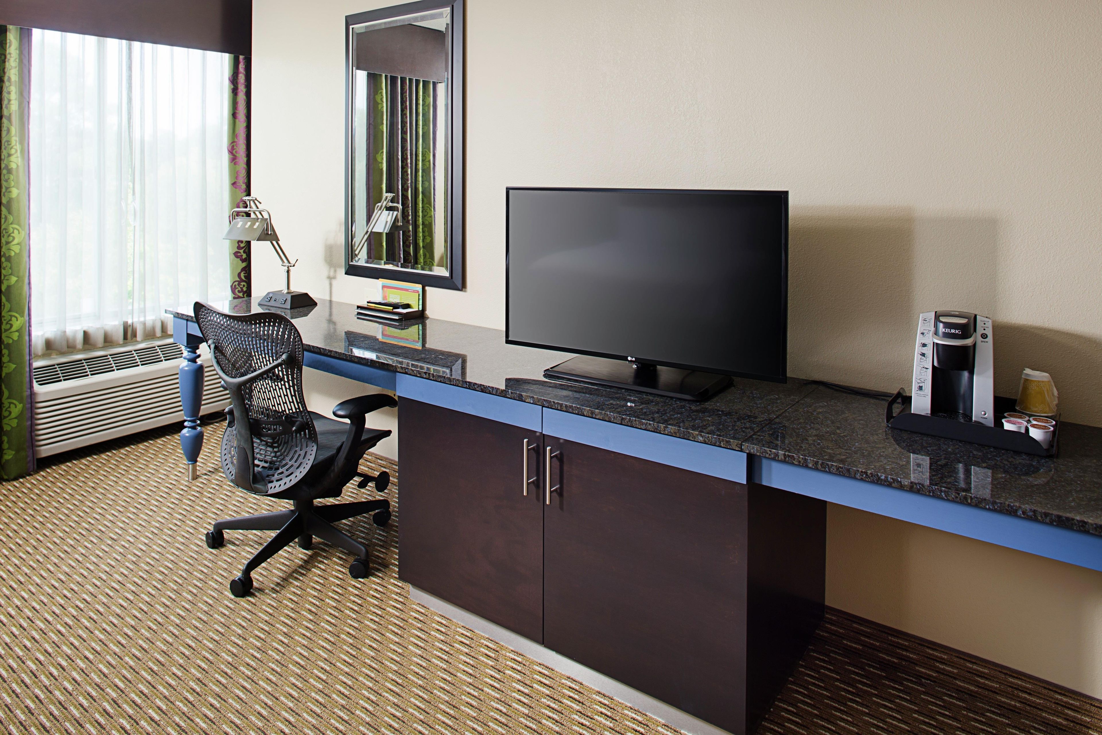A hotel room with a desk, a chair, a flat-screen TV, a coffee maker, and a window with curtains.