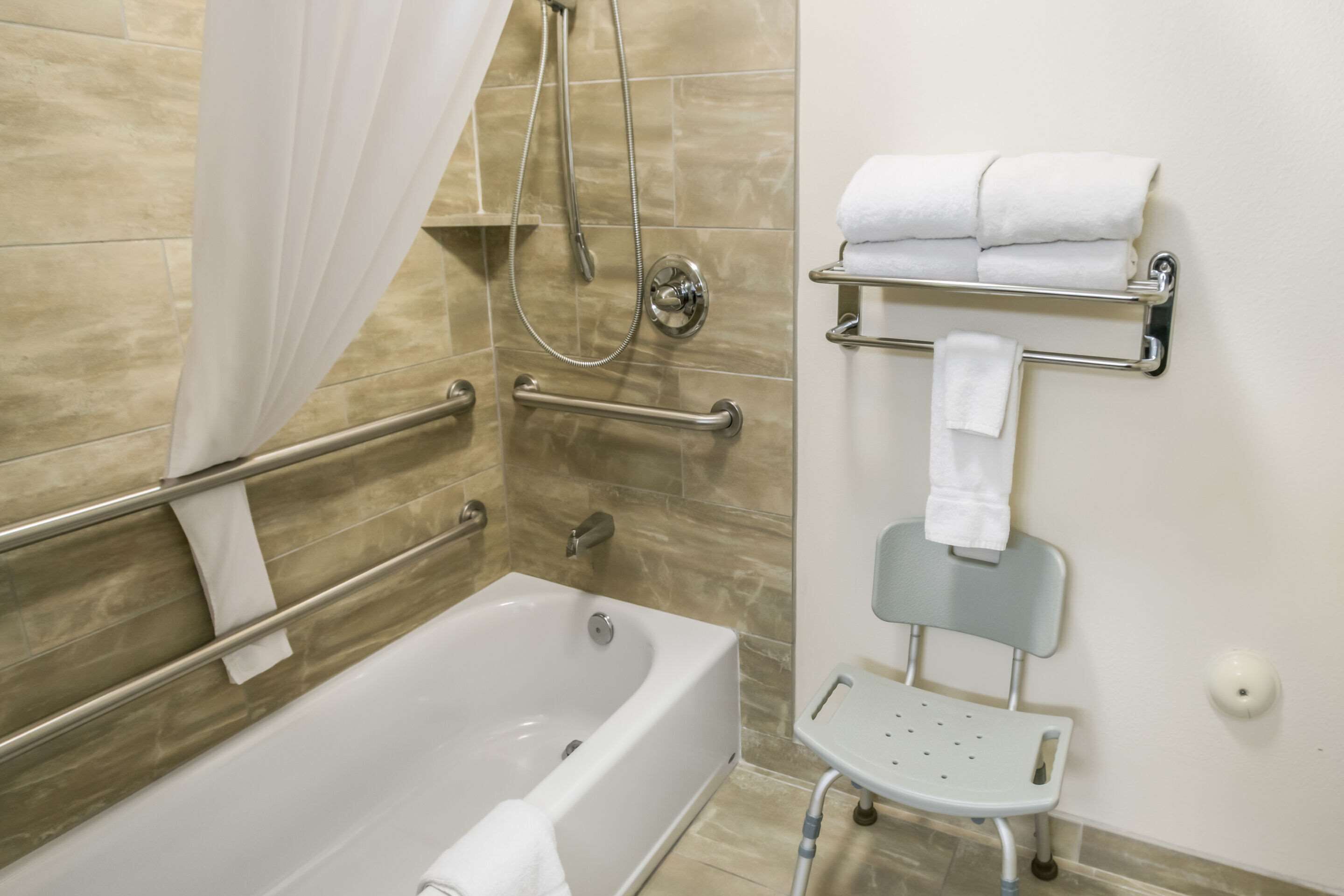 Une salle de bain équipée d’une baignoire, d’une douche avec un pommeau de douche amovible, d’une barre d’appui, d’un porte-serviettes et d’un petit tabouret.