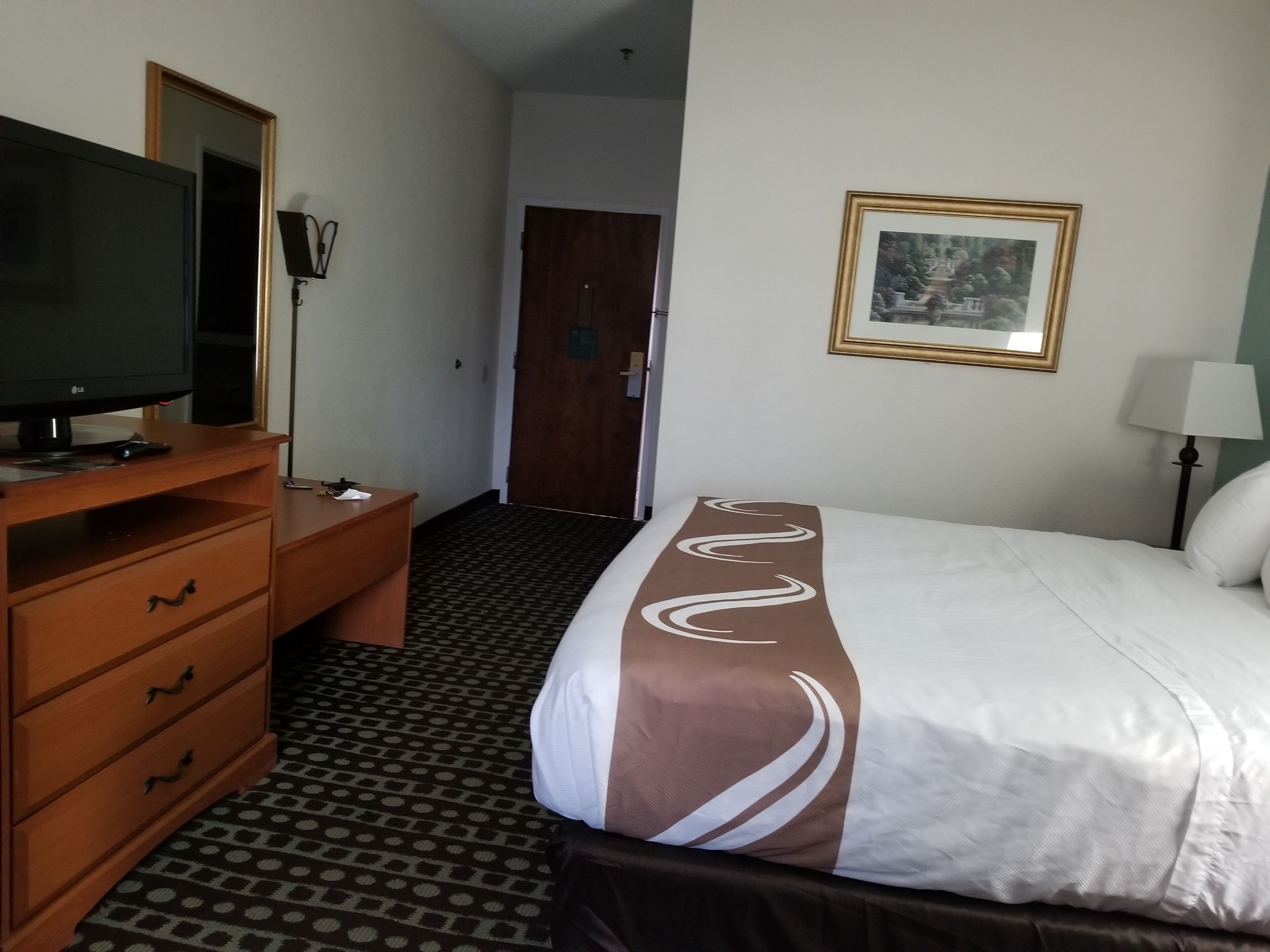 A hotel room with a bed, a TV on a wooden dresser, a framed picture on the wall, and a lamp beside the bed.