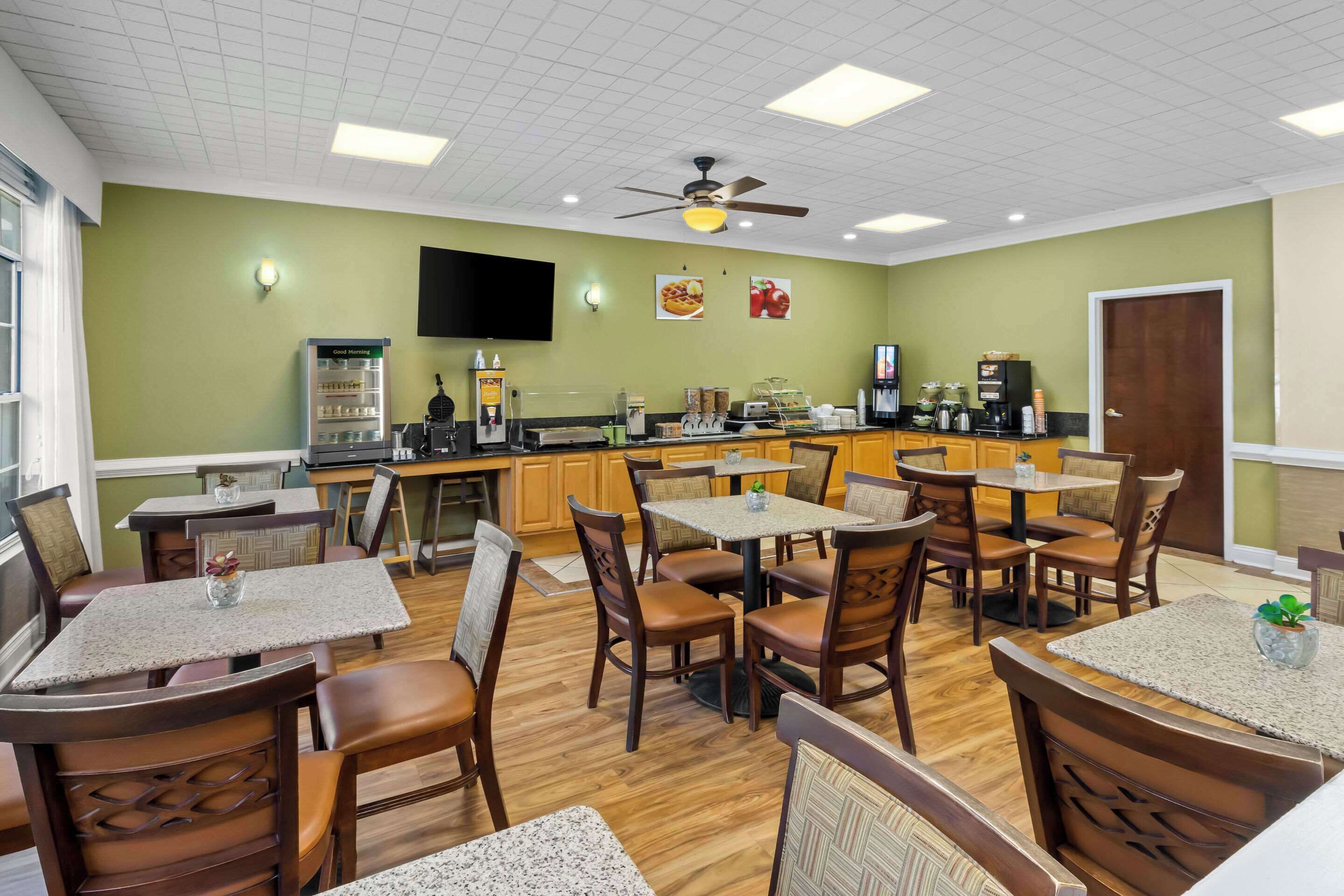 A restaurant with wooden tables and chairs, a kitchen area, and a ceiling fan.