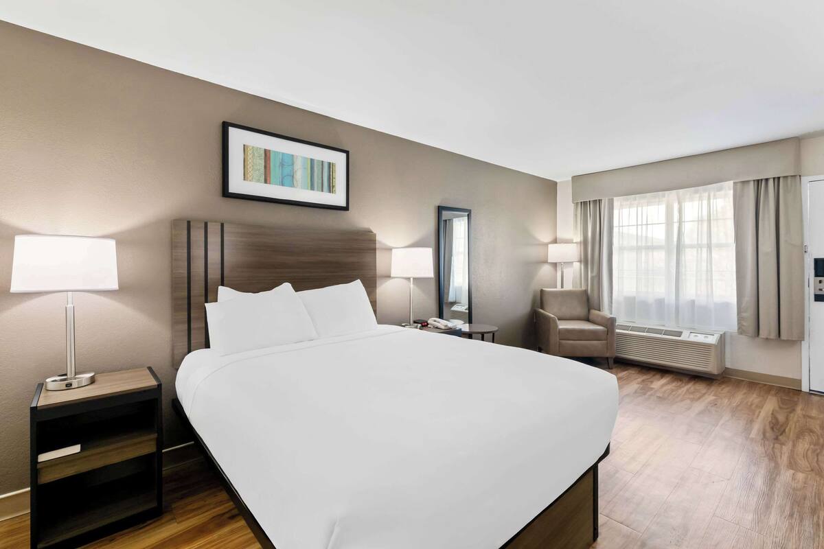 Habitación de hotel con una cama grande, lámparas de noche, una silla, un escritorio, un espejo y una ventana con cortinas.
