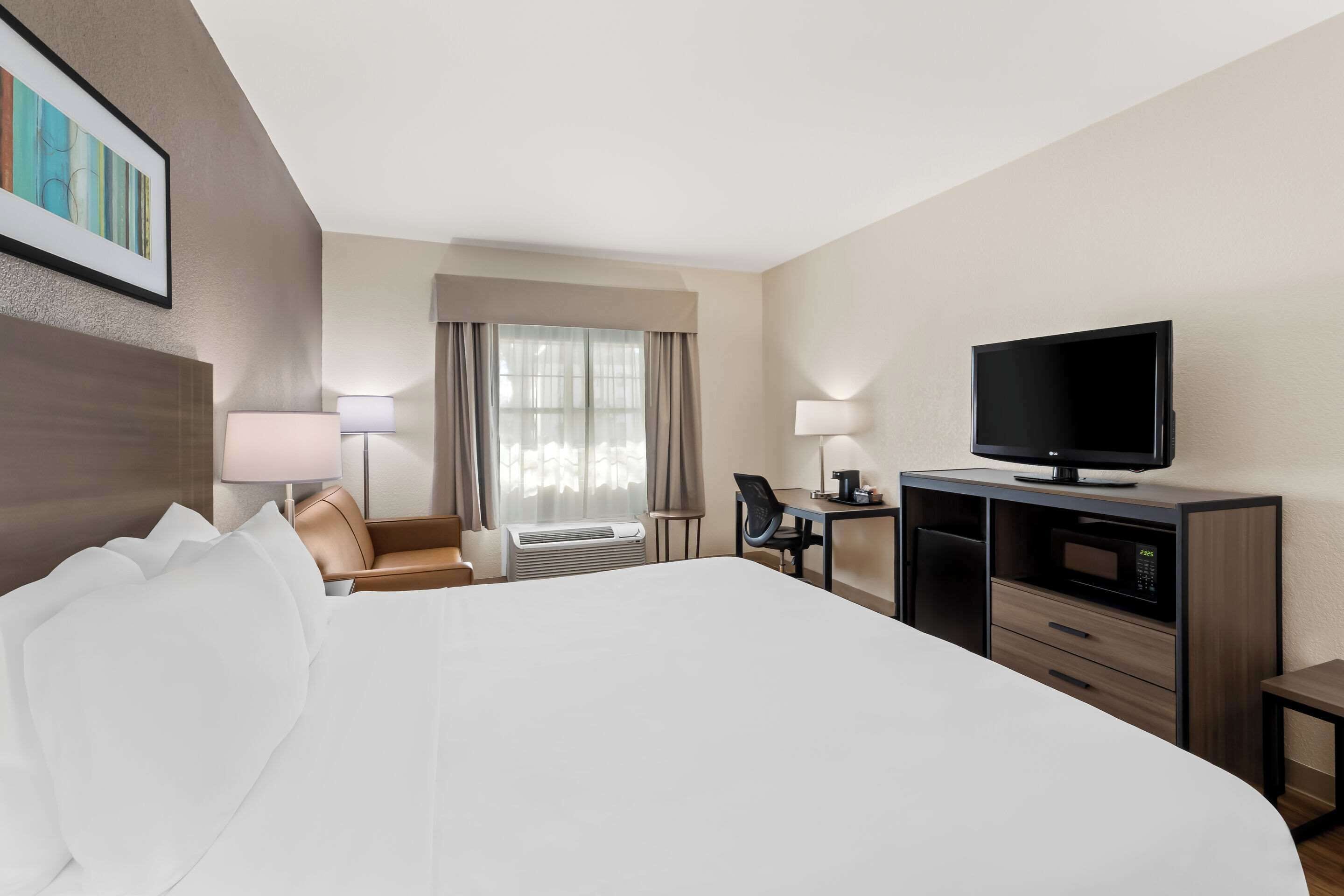 A hotel room with a large bed, a desk with a flat-screen TV, a chair, a lamp, and a window with curtains.