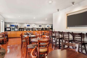 Un comedor amplio con mesas y sillas de madera, una cocina al fondo y una pantalla grande en la pared.