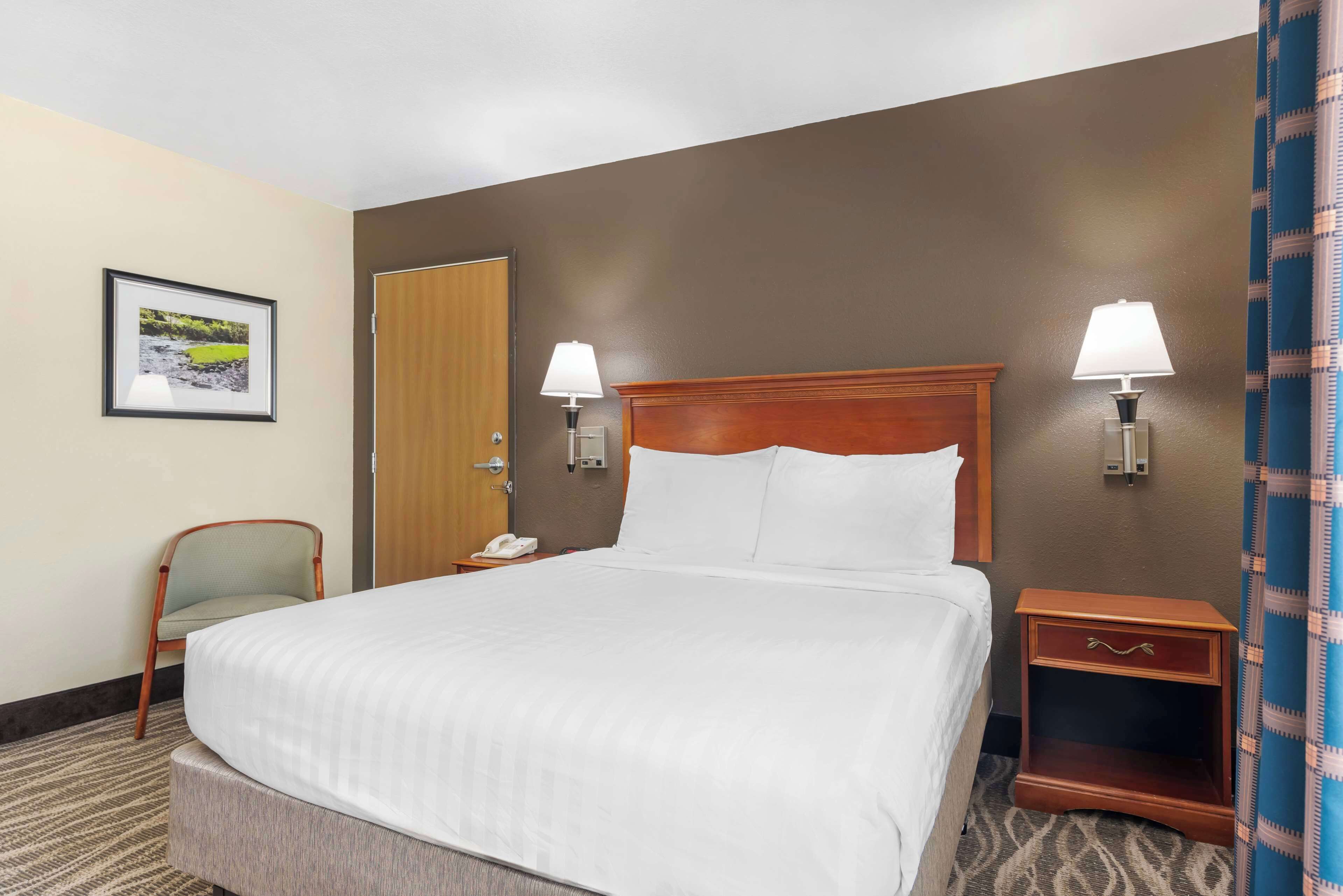 A hotel room with a large bed, two bedside lamps, a wooden nightstand, a chair, a framed picture on the wall, and a door.