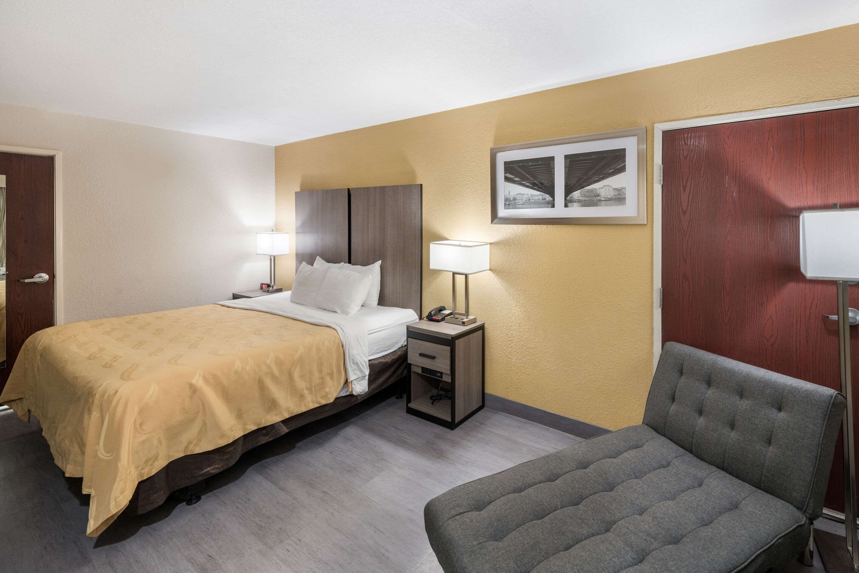 Quarto de hotel com cama, mesas de cabeceira, uma cadeira, um quadro na parede e uma porta.