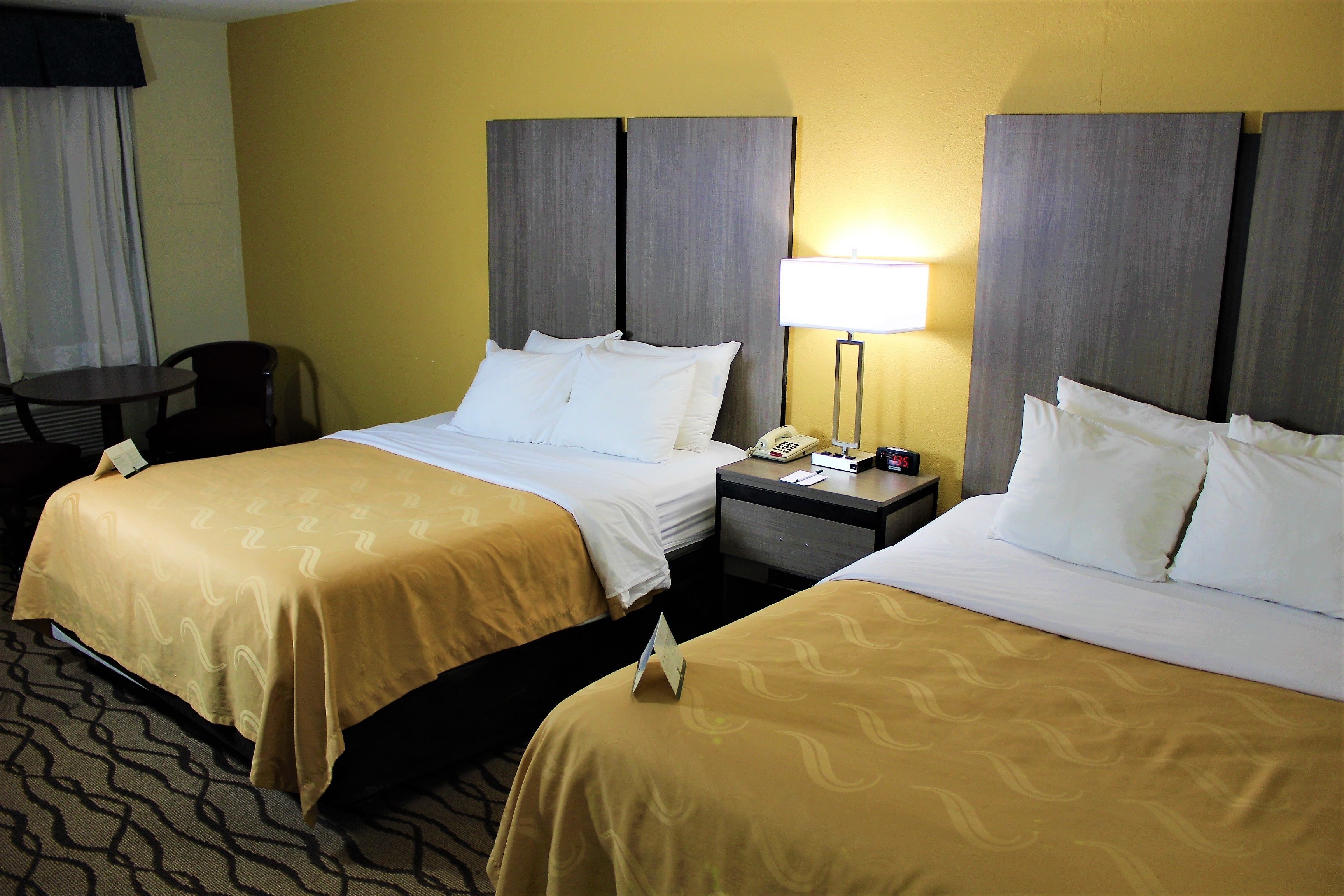 Quarto de hotel com duas camas, uma escrivaninha, uma cadeira e uma mesa de cabeceira com abajur.