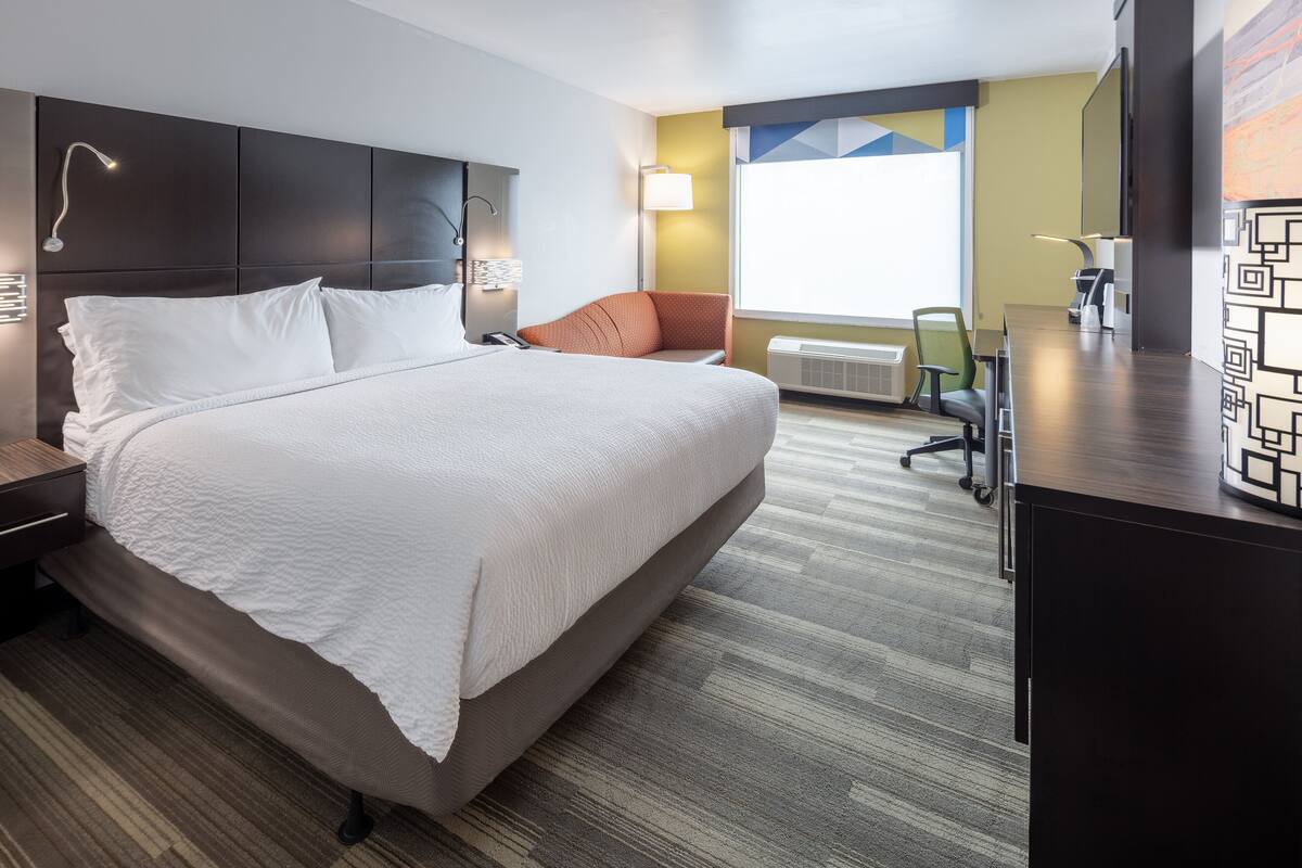 Une chambre d’hôtel moderne, dotée d’un grand lit, d’un bureau et d’une chaise. Une fenêtre laisse entrer la lumière naturelle et offre une vue sur l’extérieur.