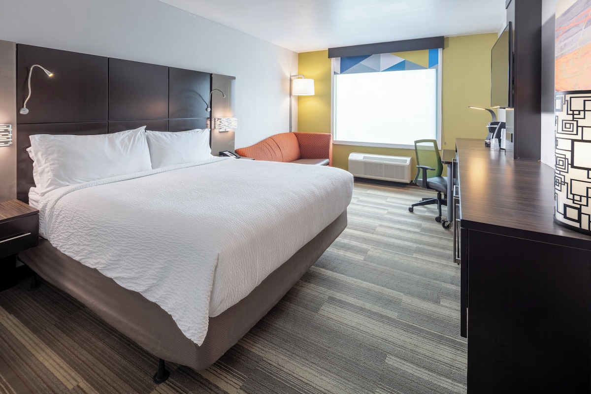 A modern hotel room with a large bed, a desk, and a chair. There is a window providing natural light and a view of the outside.