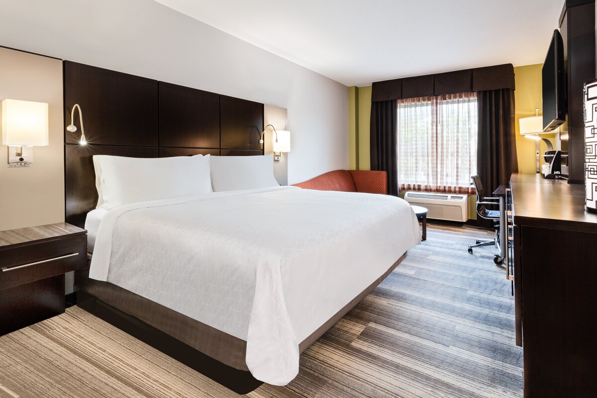 Une chambre d’hôtel avec un grand lit, un bureau et une chaise. Il y a deux lampes sur le mur et un téléviseur posé sur un meuble.