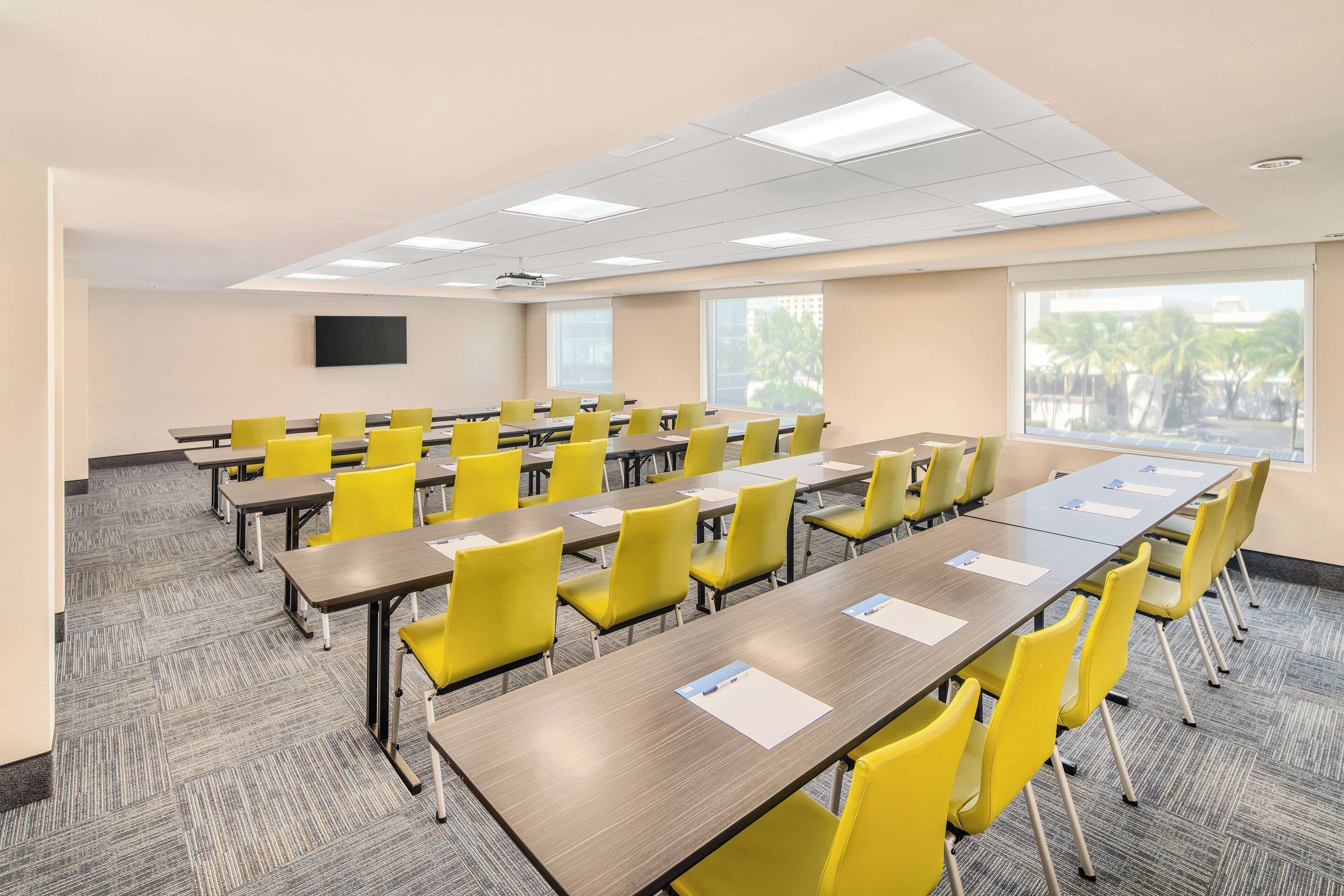 Une salle de conférence équipée de plusieurs tables et chaises, de grandes fenêtres et d’un téléviseur à écran plat fixé au mur.