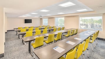 Un salón de conferencias con varias mesas y sillas, ventanas amplias y un televisor de pantalla plana en la pared.
