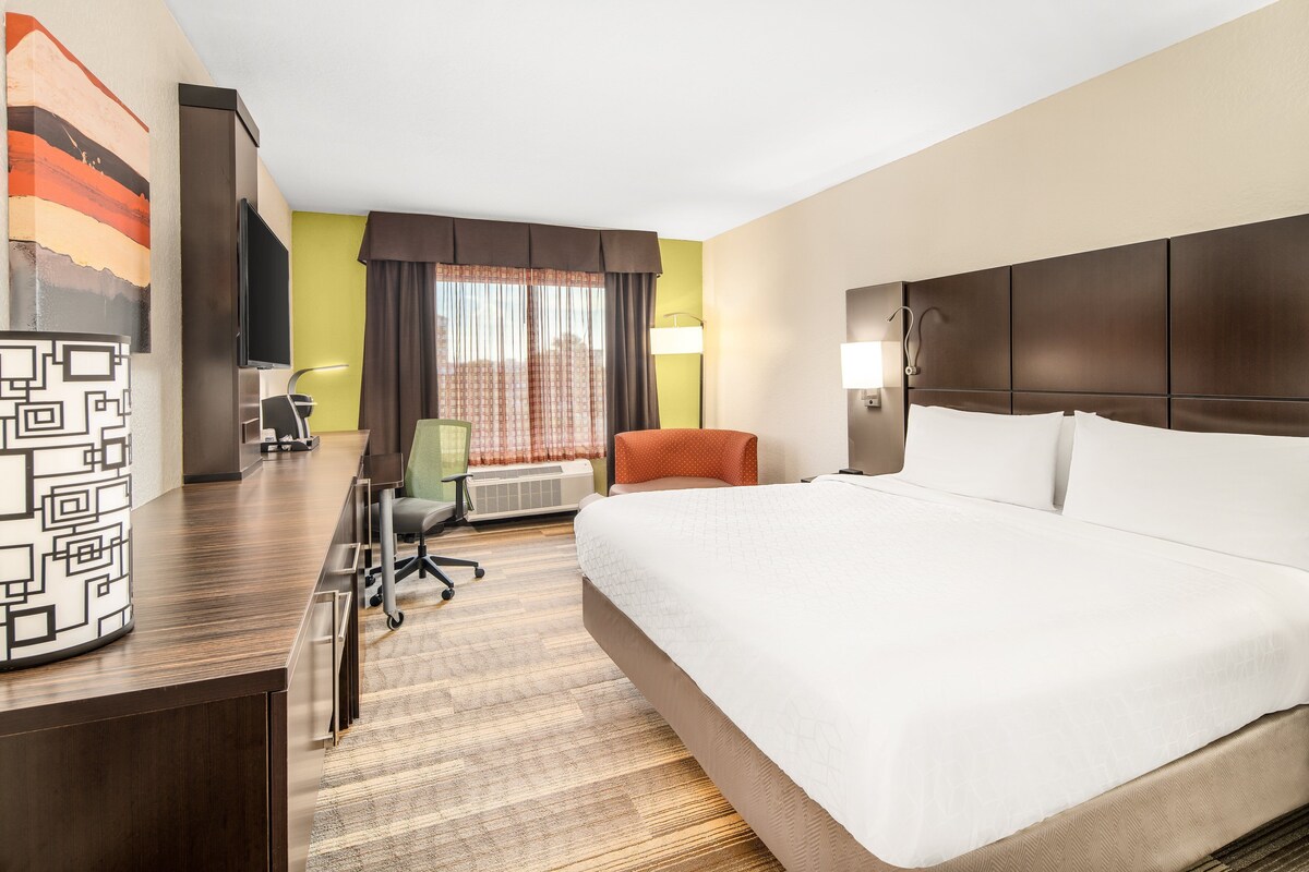 Une chambre d’hôtel avec un grand lit, un bureau avec un ordinateur, une chaise et une fenêtre avec des rideaux.