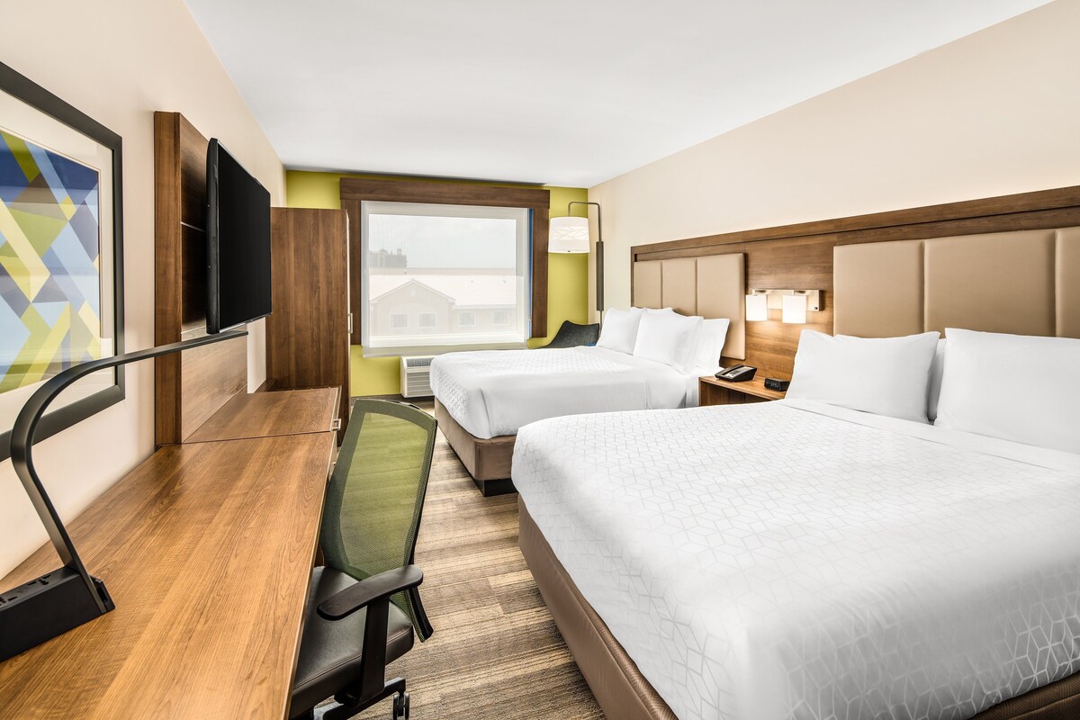 Une chambre d’hôtel avec deux lits, un bureau, une chaise, un téléviseur à écran plat et une fenêtre donnant sur des immeubles.
