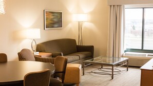 A hotel room with a sofa, armchairs, a glass-top coffee table, a dining table, and lamps.