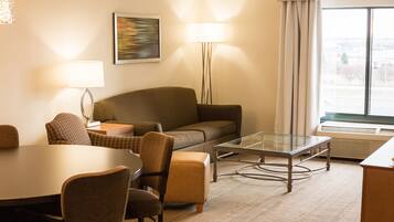 Habitación de hotel con sofá, sillones, una mesa de centro de vidrio, una mesa de comedor y lámparas.