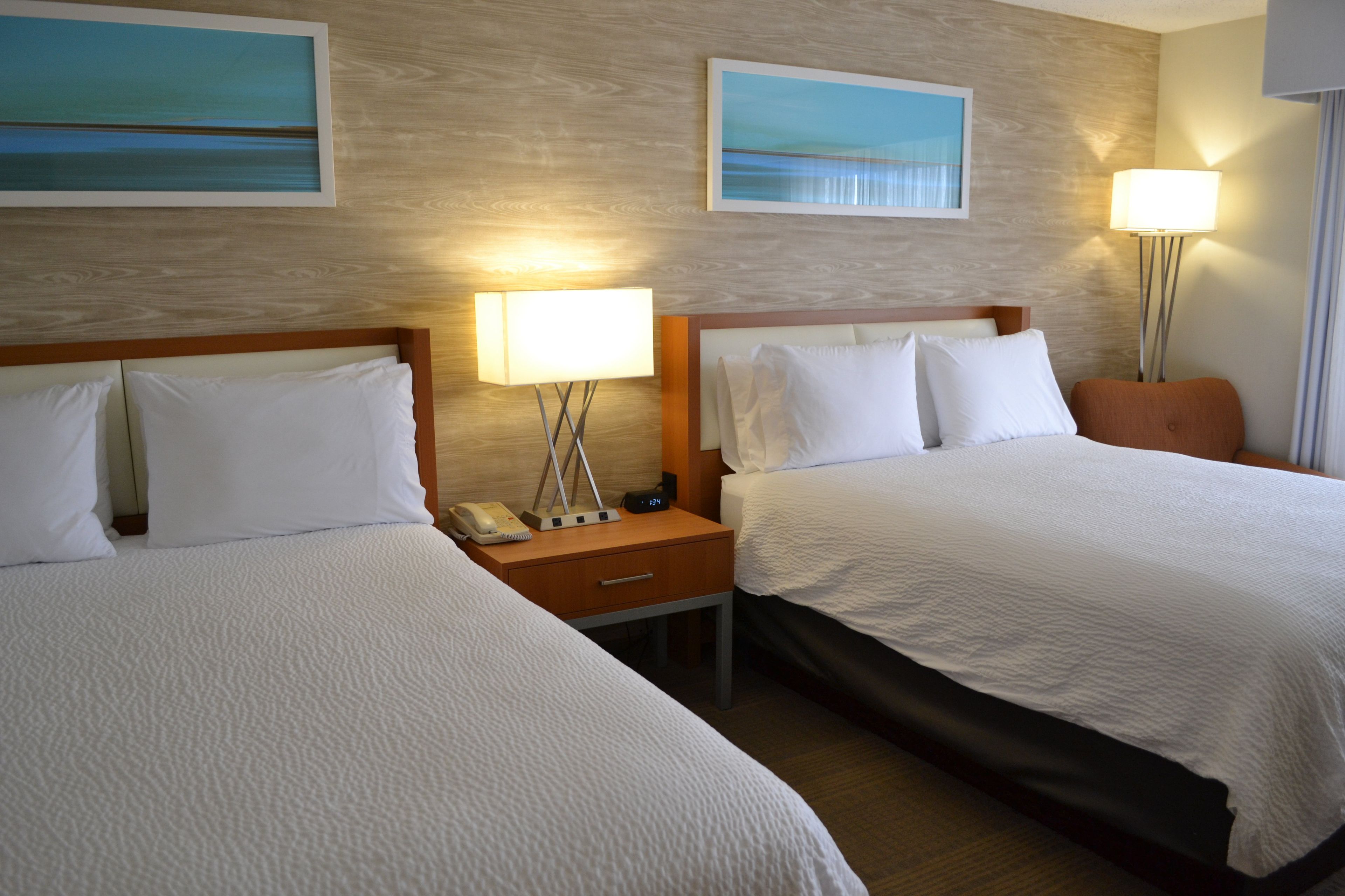 A hotel room with two beds, a nightstand with a lamp, a phone, and a digital clock.