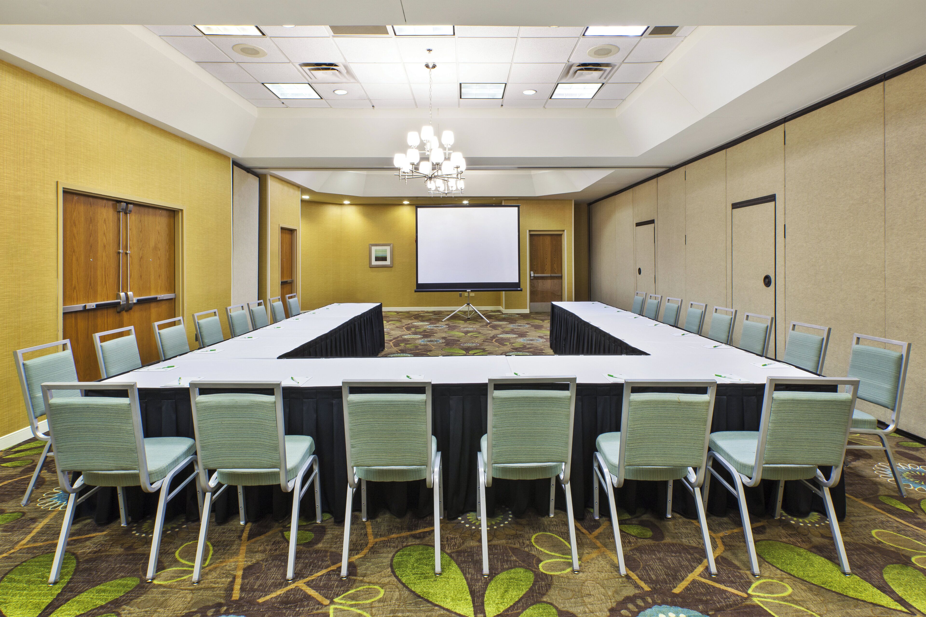 A conference room with a long table, chairs, a projector screen, and a carpeted floor.