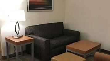 A hotel room with a sofa, two ottomans, a small table with a lamp, and a painting on the wall.