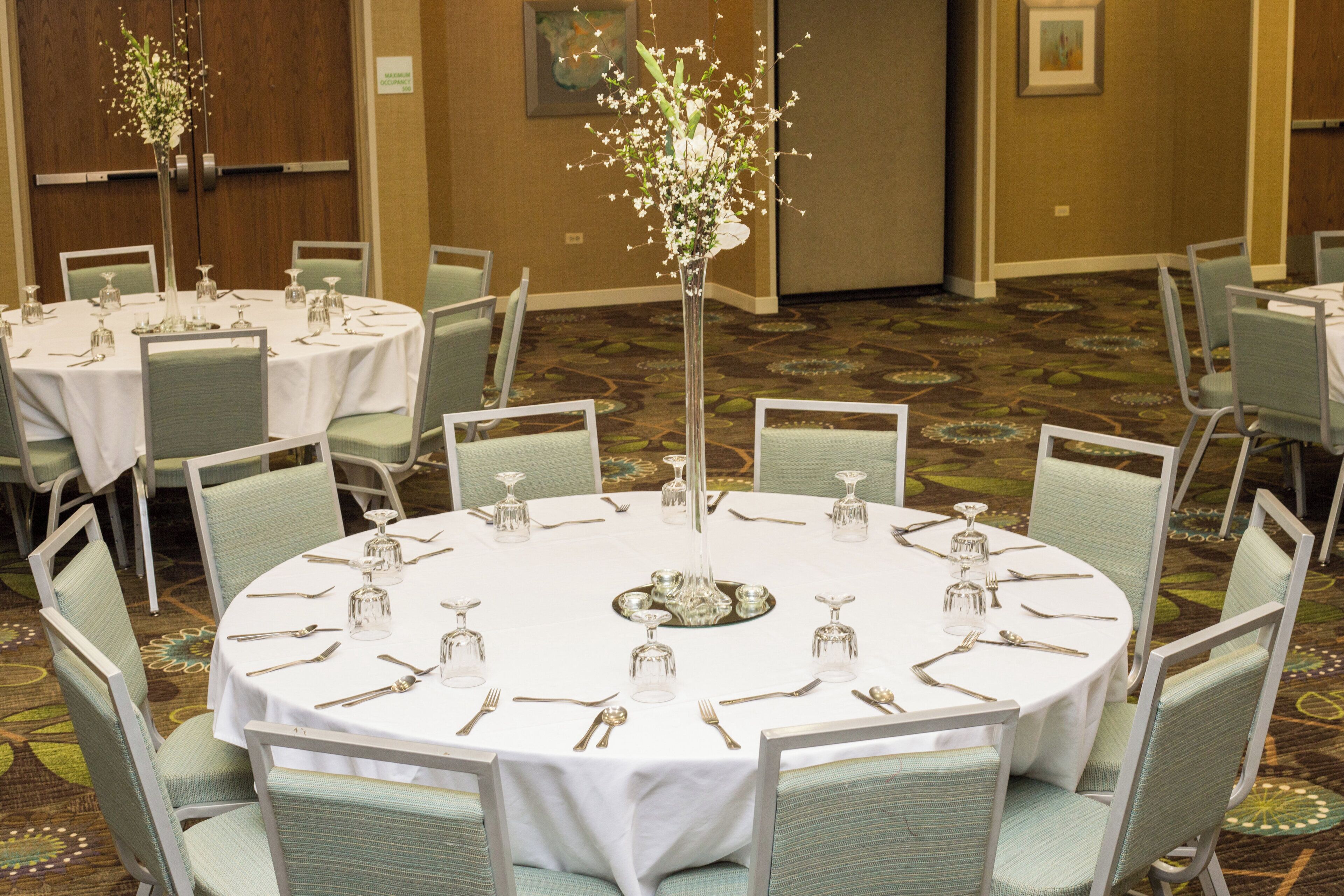 A banquet hall with round tables set for dining, chairs, and a central floral arrangement.