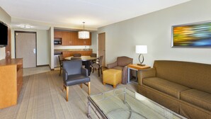 A hotel room with a living area, a glass coffee table, a sofa, a TV, a desk, a chair, a lamp, and a painting on the wall.