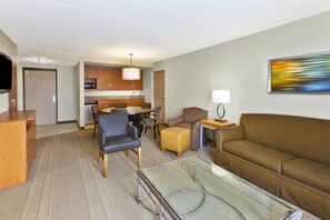 A hotel room with a living area, a glass coffee table, a sofa, a TV, a desk, a chair, a lamp, and a painting on the wall. - Holiday Inn & Suites Bolingbrook by IHG (Bolingbrook)