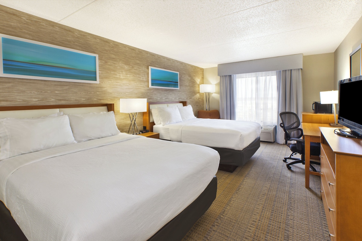 A hotel room with two beds, a desk, a chair, a television, and artwork on the walls.