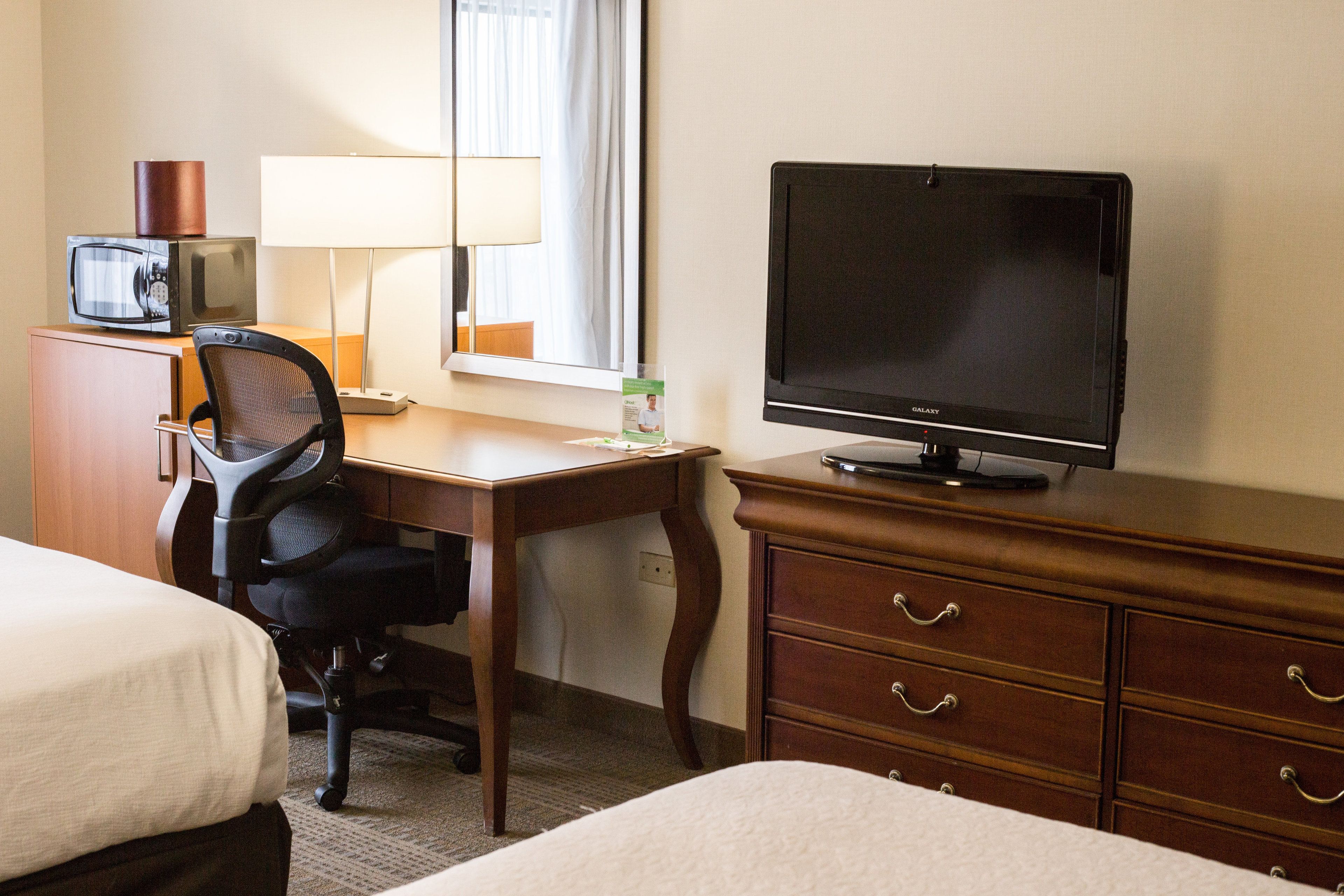 Une chambre d’hôtel équipée d’un lit, d’un bureau avec un ordinateur, d’une télévision, d’une lampe et d’un four à micro-ondes.