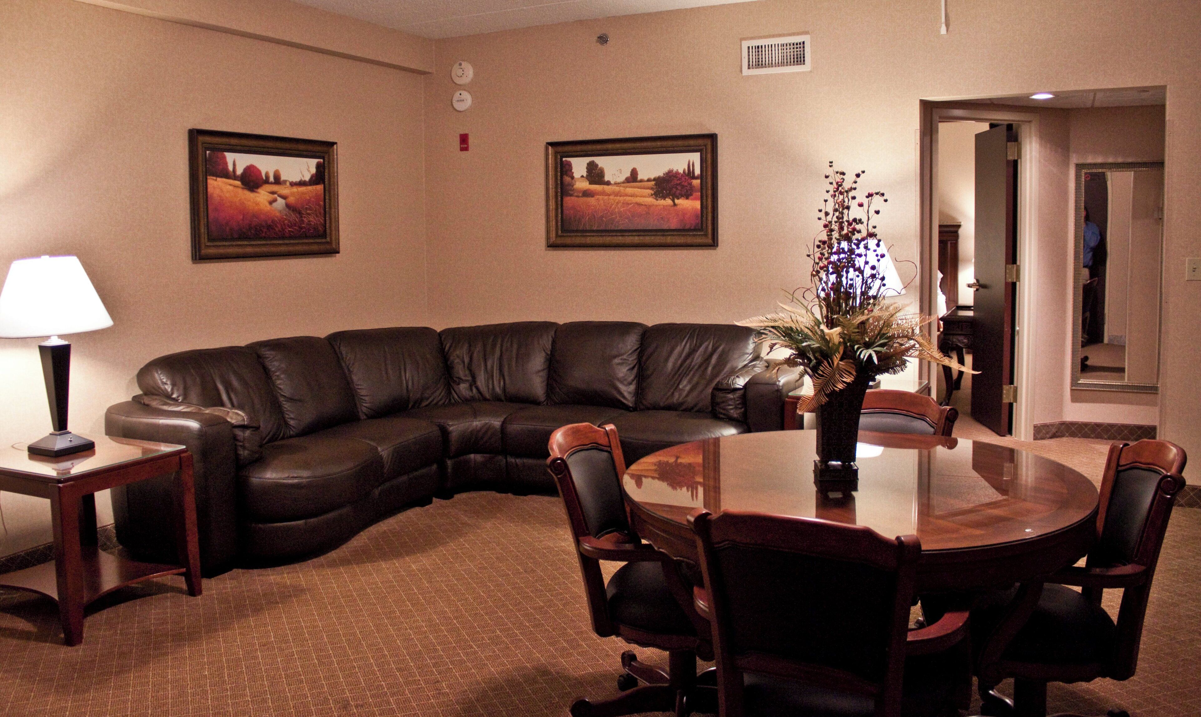 Ein Hotelzimmer mit einer gebogenen Ledersofa, einem Esstisch mit Stühlen, einer Vase mit Blumen und zwei gerahmten Bildern an der Wand.