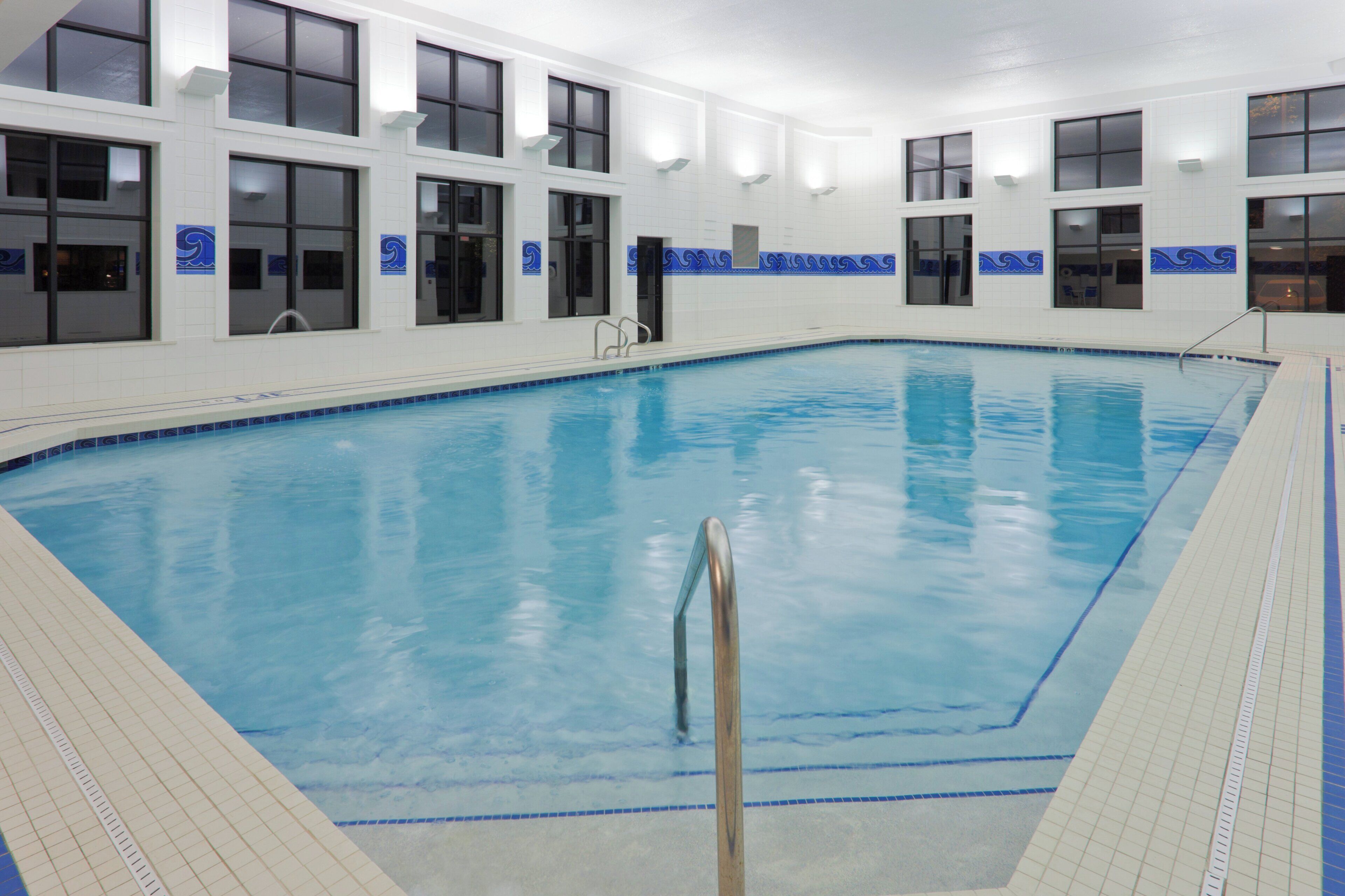 An indoor swimming pool with clear blue water, surrounded by white tiled walls with blue decorative patterns, and large windows.