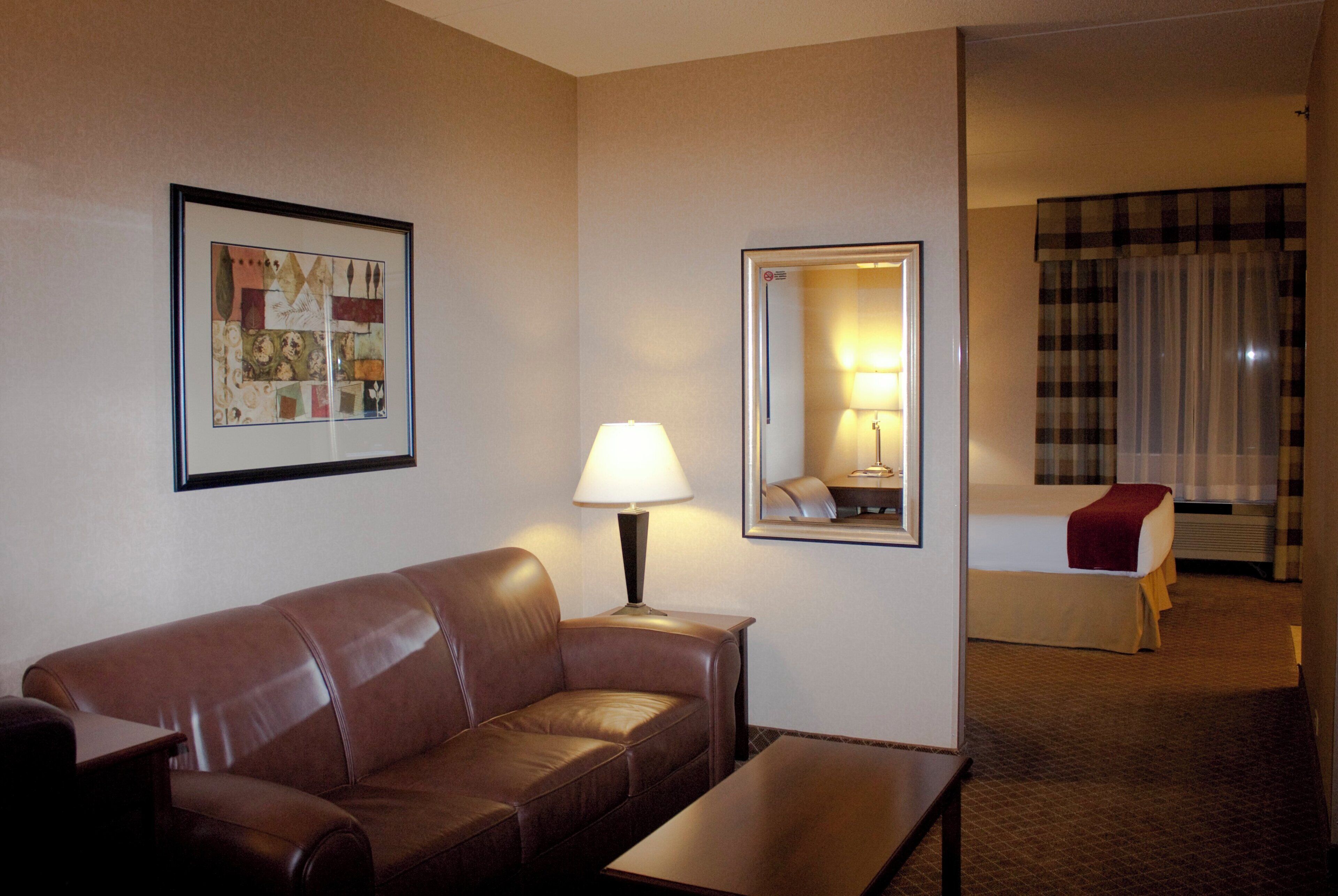 A hotel room with a brown leather sofa, a bedside table, a lamp, a bed with a red blanket, a mirror, and a framed picture on the wall.