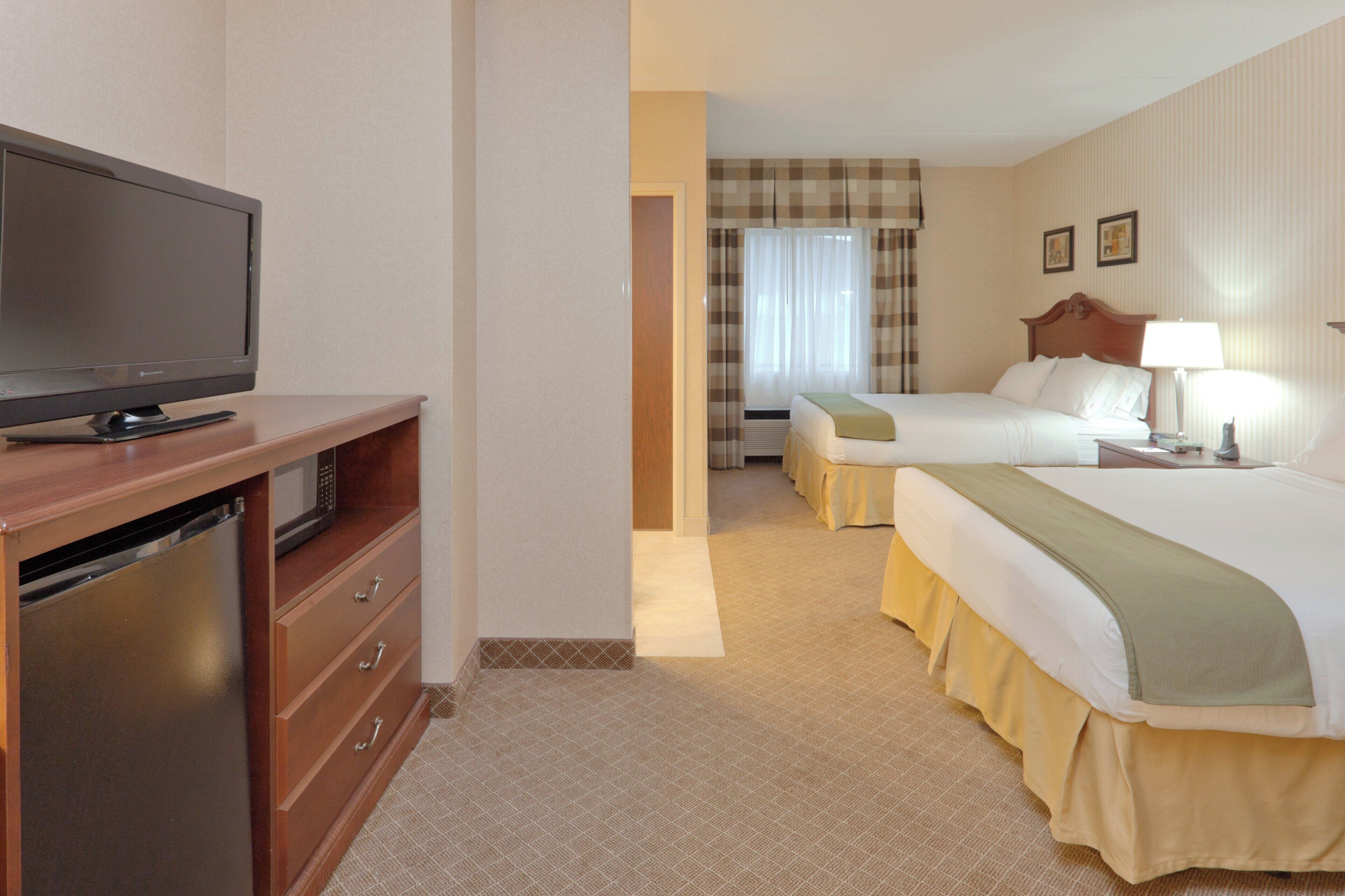 A hotel room with a television on a wooden cabinet, a bed with a green throw, a bedside table with a lamp, and a checkered curtain.