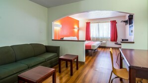 Une chambre d’hôtel avec un canapé vert, une table en bois, un petit tabouret en bois, un lit recouvert d’une couverture rouge, un bureau avec une chaise et un miroir qui reflète la pièce.