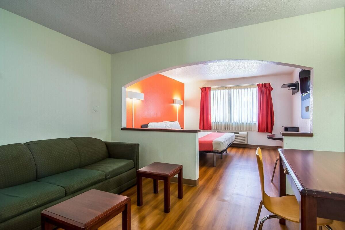 A hotel room with a green sofa, a wooden table, a small wooden stool, a bed with a red blanket, a desk with a chair, and a mirror reflecting the room.