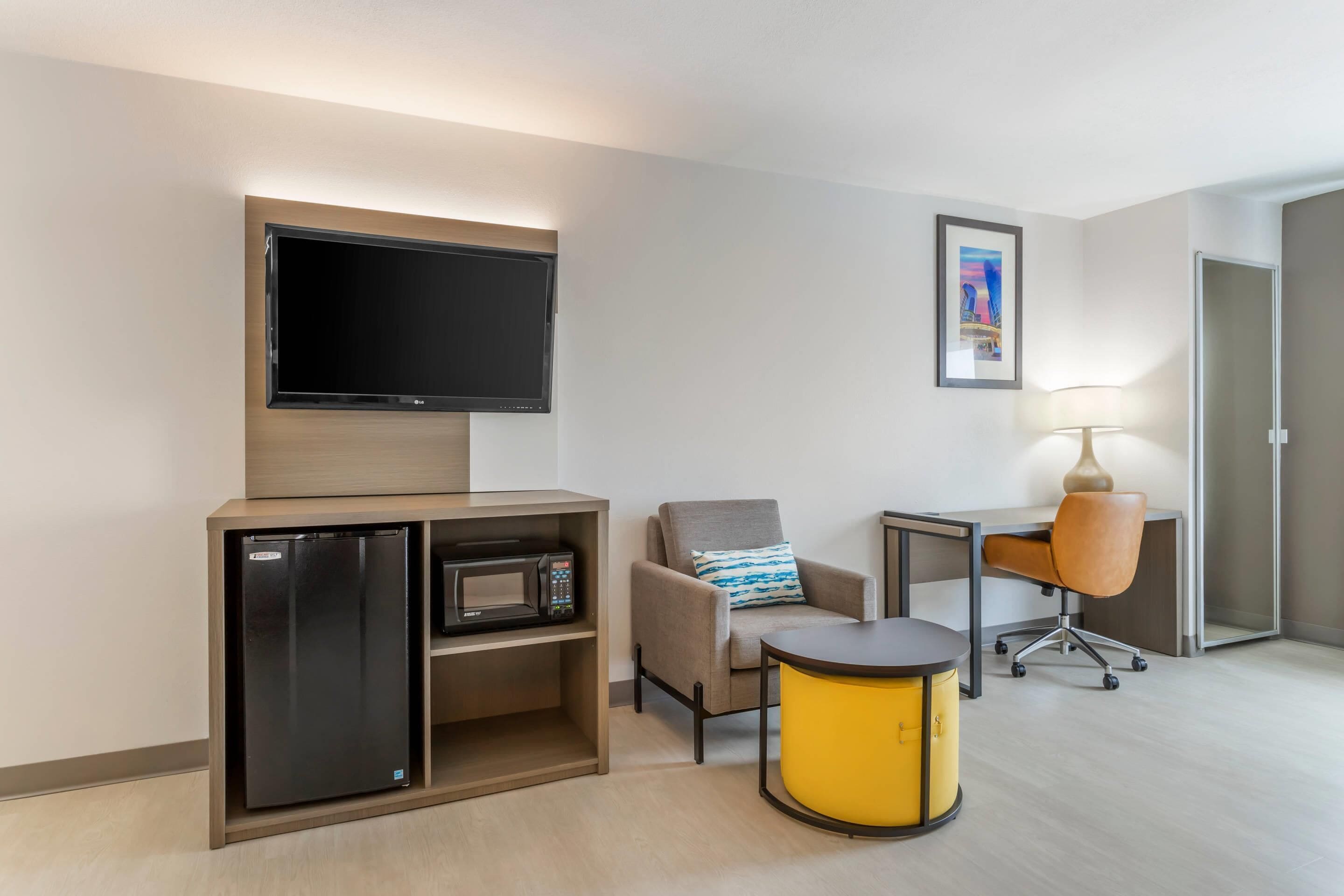 Een hotelkamer met een flatscreen televisie boven een minibar, een comfortabele fauteuil, een bureau met lamp en een klein rond tafeltje.