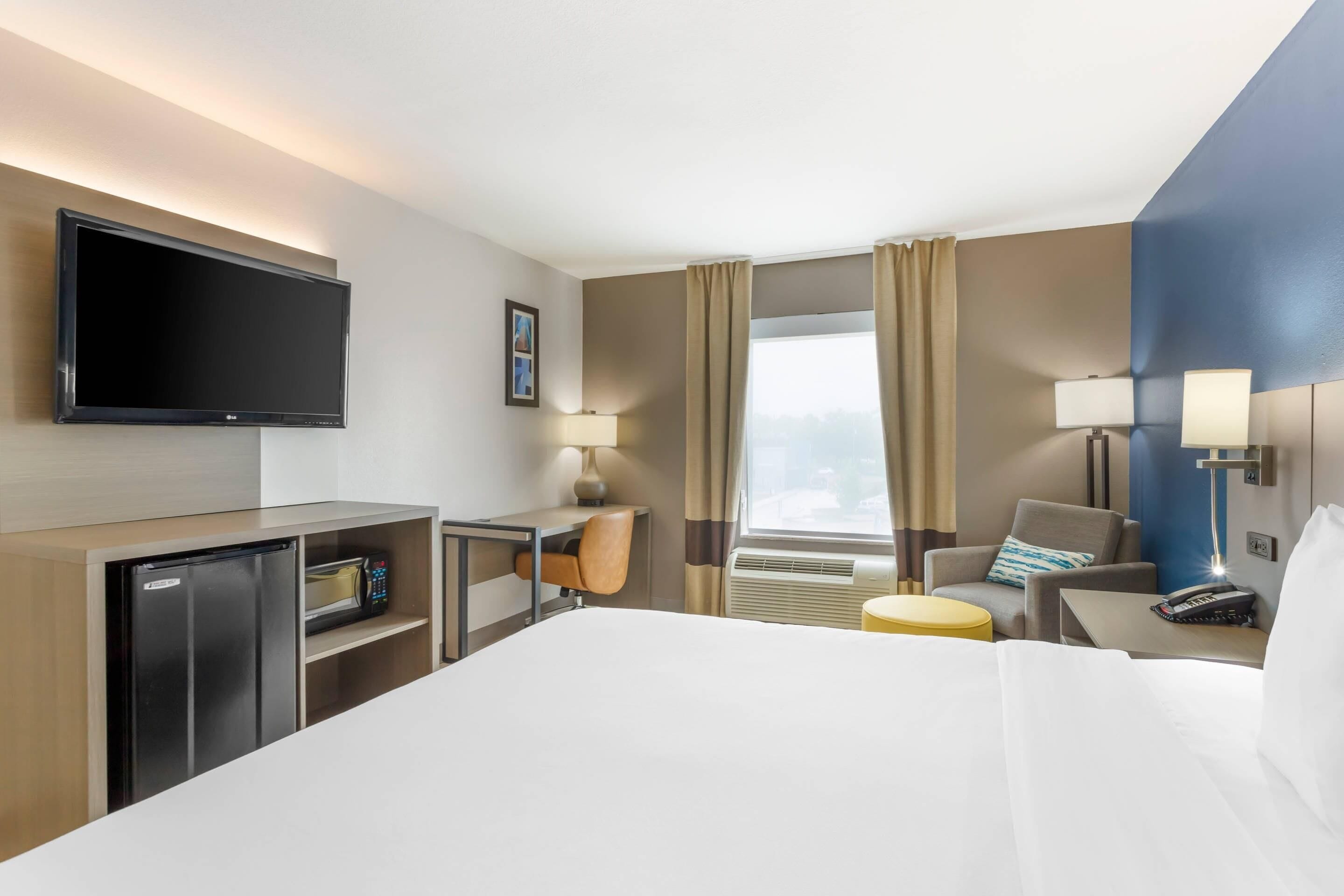 Een hotelkamer met een bed, een bureau met computer, een televisie, een magnetron, een stoel, een lamp en een raam met gordijnen.