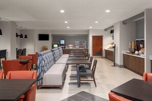 A modern dining area with a long bench, individual tables, and chairs. There is a counter with food items and a television mounted on the wall. - Holiday Inn Express Greenville by IHG (Greenville)